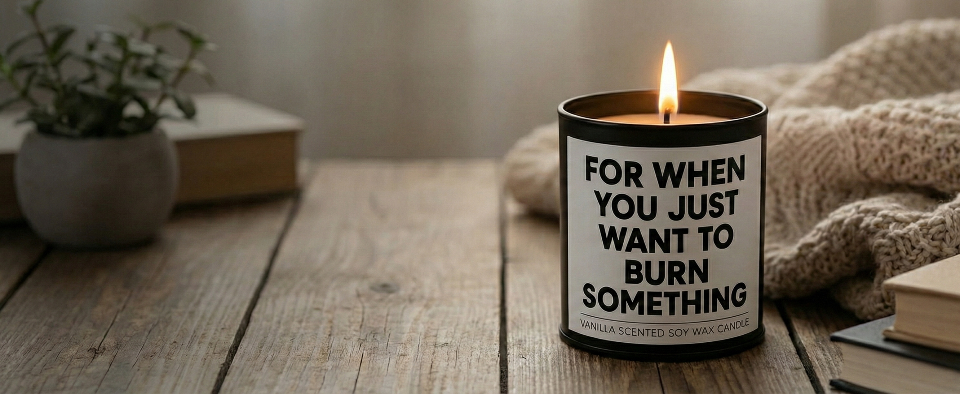 Candle with a humorous label on a wooden surface, surrounded by books and a plant.
