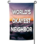 A rectangular garden flag with a purple and orange cloud-filled sky background and the text 'WORLD'S OKAYEST NEIGHBOR' in bold white letters.